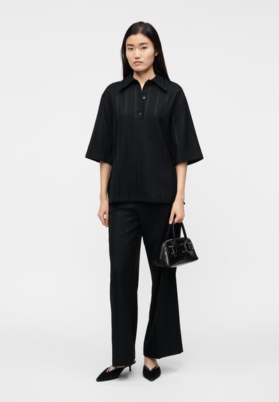 Camicia nera a maniche corte con bottoni e trama a righe, abbinata a pantaloni neri a righe sottili e scarpe con punta appuntita, con una piccola borsa nera in mano.