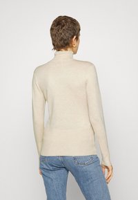 Beige Rollkragenpullover aus weichem Material, mit langen Ärmeln und figurbetontem Design. Kombiniert mit hellblauen Jeans.