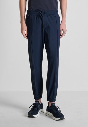 Hombre con pantalones jogger azul marino con puños elásticos y zapatos deportivos negros, de pie sobre un fondo claro y simple.