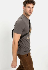 Graues Baumwoll-T-Shirt mit einem grafischen Design auf der Vorderseite, kombiniert mit braunen Cordhosen. Modell trägt eine schwarze Umhängetasche.