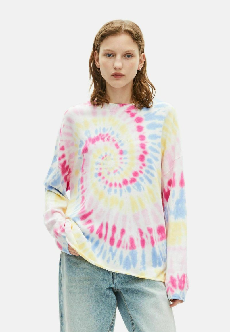 Jonge vrouw in een pastel tie-dye langemouwen shirt en lichtblauwe jeans, staand met één hand in de zak, neutrale achtergrond.