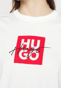 Camiseta de algodón blanca con un logo cuadrado rojo que lleva "HUGO" en letras blancas en negrita y un acento en cursiva negra. Cuello redondo, mangas cortas.