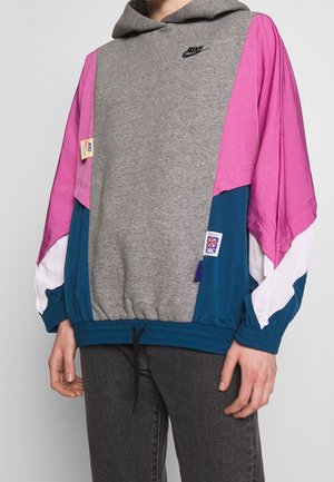 Sweat à capuche Nike avec devant gris, manches roses, panneaux bleus, accents blancs, logo Nike noir sur la poitrine et patchs sur les manches.