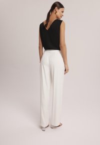 Czarny bezrękawnik z dekoltem w kształcie litery V, połączony z białymi spodniami o wysokim stanie, charakteryzującymi się gładką fakturą i plisowanymi detalami. Widoczne przezroczyste obcasy.