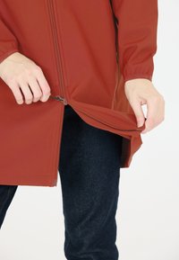 Punainen synteettinen takki, jossa on vetoketjullinen etuosa, osittain auki. Mallilla on päällään tummansiniset farkut. Takissa on suora leikkaus ja pitkät hihat.