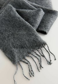 Szary wełniany szalik o miękkiej, puszystej teksturze z frędzlami na końcach. Materiał wydaje się ciepły i przytulny.