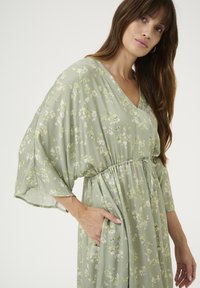 Obleka svetlo zelene barve s cvetličnim potiskom, z V-izrezom, ohlapnimi rokavi v obliki netopirja, elastiko v pasu in stranskimi žepi, narejena iz padajočega materiala.