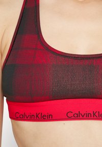 Červená a čierna károvaná športová podprsenka so širokým červeným pásom s logom "Calvin Klein", vyrobená z elastického, textúrovaného materiálu.