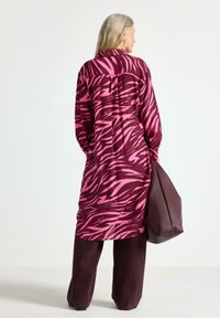 Lang skjorte i et pink og burgunderrandet zebra print, lavet af glat stof. Sammen med mørke, vide bukser og en stor brun taske.