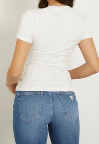 Dopasowana biała koszulka bawełniana z krótkim rękawem, w zestawieniu z niebieskimi dżinsami o klasycznym pięciopocketowym wzorze oraz trójkątnym logo na tylnej kieszeni.