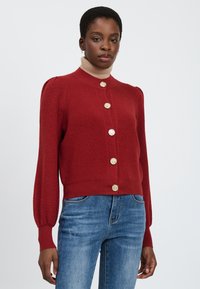 Rode gebreide cardigan met een hoge kraag, zeven decoratieve knopen en poflange mouwen. Draagt over een beige coltrui en gecombineerd met blauwe spijkerbroek.