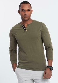 Mann in einem olivgrünen Langarm-Henley-Shirt mit Holzknöpfen und hellen Hosen, stehend mit einer Hand in der Tasche.