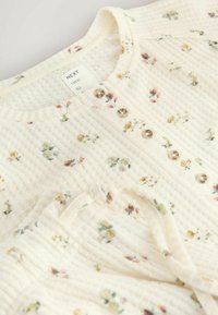 Hvid ribstrikket stoftop med små blomsterprint, forsynet med fem gennemsigtige knapper langs fronten og med rund halsudskæring.