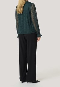 Haut à manches longues en dentelle turquoise avec motifs géométriques, associé à un pantalon noir à jambes larges et des chaussures pointues noires. Texture lisse et coupe ample.