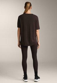 Kobieta z kucykiem, ubrana w czarną, oversizową koszulkę, czarne legginsy, czarne skarpetki i czarne sneakersy, stoi tyłem na neutralnym tle.