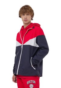 Giacca impermeabile bicolore con blocchi di colore rosso, bianco e blu navy, dotata di chiusura con zip, cappuccio regolabile e tasche laterali per una maggiore praticità.