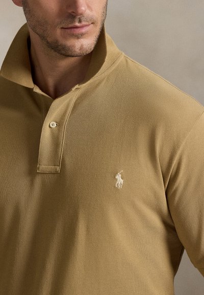 Khaki polo shirt with a textured fabric, short collar, three-button placket, and an embroidered white logo on the left chest.