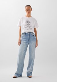 Witte oversized T-shirt met een schedelgrafiek en tekst. Lichtblauwe wijde jeans. Gladde stof, casual pasvorm, minimalistisch ontwerp.