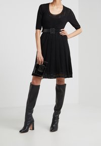 Abito nero in maglia con maniche corte, vita aderente e gonna plissettata. Abbinato a stivali neri con tacco alti fino al ginocchio, una cintura nera e una pochette nera.
