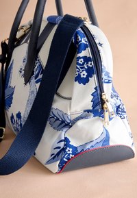 Blå och vit blommig canvas-handväska med guldfärgad dragkedja, texturerade marinblå remmar och en grå botten med en röd accent.