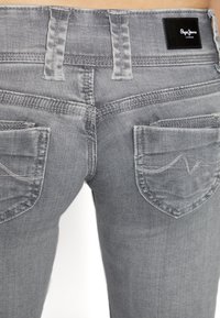 Jeans de mezclilla gris con bolsillos traseros y pasadores para cinturón, llevados por una persona, con una etiqueta negra de Pepe Jeans London situada encima del bolsillo derecho.