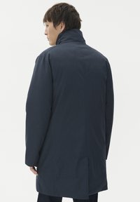 Jūras zila mētelis ar augstu apkakli, strukturēta auduma, garām piedurknēm un taisnu griezumu. Iezīmē aizmugurē esošo atvērumu kustīguma nodrošināšanai un minimālistisku dizainu.