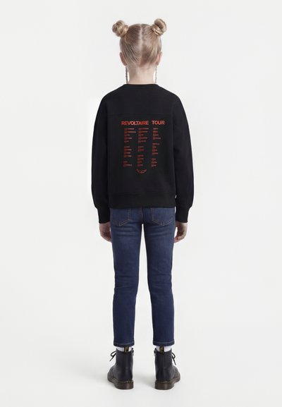 Kind mit blondem Haar in zwei Dutts trägt schwarzen Pullover mit roten Städtenamen, kombiniert mit blauen Jeans und schwarzen Stiefeln, steht mit dem Rücken zur Kamera.