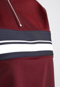 Burgundská horní látka s polovičním zipem, doplněná pruhovaným námořnickým a bílým páskem přes oblast hrudníku. Hladká textura.