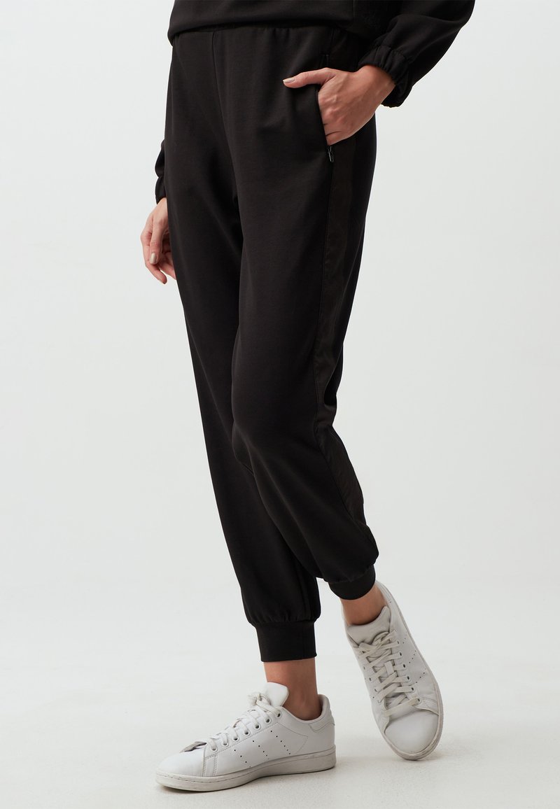 Zwarte joggingbroek van zacht materiaal met elastische boorden en zijzakken, gecombineerd met witte sneakers. Soepele textuur, eenvoudig ontwerp.