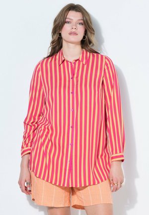 Kvinde iført lyserød, gul og orange lodret striped knap-op skjorte med ferskenfarvede shorts mod en ensfarvet lys baggrund.