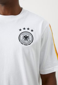 Hvit bomull t-skjorte med en svart brodert Tyskland Fußball-Bund-logo med fem stjerner over. Røde og gule striper på ermene.