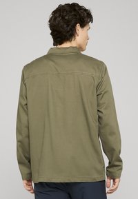 Chemise à manches longues vert olive avec col, coutures visibles et tissu lisse et texturé. Vue de dos montrant un design simple et une coupe décontractée.