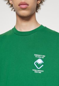 Camiseta verde de algodón con cuello redondo, mangas cortas y un logo bordado con detalles de montaña y texto en la parte superior izquierda.