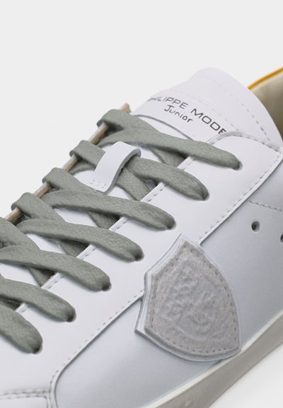 Baskets en cuir blanc avec des lacets gris, un écusson en forme de bouclier sur le côté, et le texte « Philippe Model Junior » sur la languette.