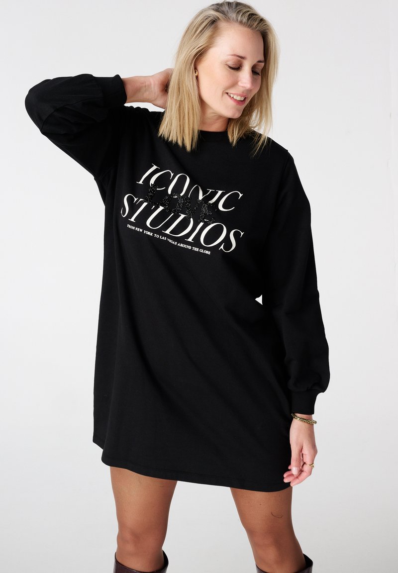 Zwart oversized sweatshirtjurk met lange mouwen, voorzien van gedurfde witte tekst met de woorden "ICONIC STUDIOS" en een zachte stoftextuur.