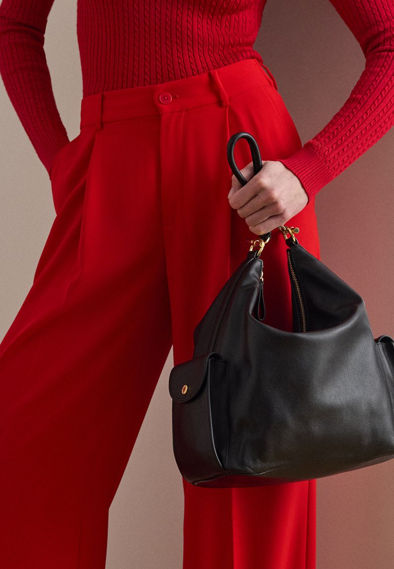 Osoba u teksturiranom crvenom džemperu i crvenim hlačama, držeći crnu kožnu torbu s zlatnim okovom za ručke.