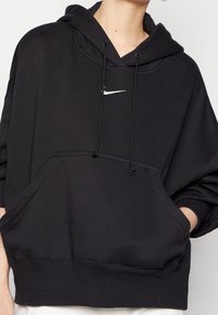 Hoodie noir avec poche kangourou à l'avant, capuche avec cordon de serrage et petit logo Nike en forme de swoosh blanc sur la poitrine. Texture de tissu douce.