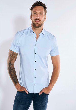 Helles blaues Kurzarmhemd mit einem geknöpften Kragen und schwarzen Knöpfen. Der Stoff wirkt glatt mit einem leichten Glanz und wird mit dunklen Jeans getragen.