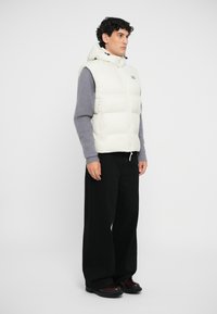 White puffer vest with a fitted hood, zippers, and down filling, layered over a gray sweater. Black wide-leg trousers and brown boots complete the look.
