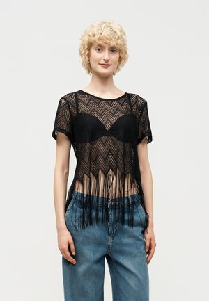 Rövid, göndör szőke hajú személy fekete áttetsző, bojtos felsőt visel fekete melltartó fölött és laza kék farmert, egyszínű háttér előtt áll.