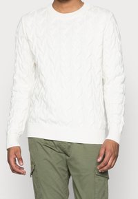 Vit kabelstickad tröja med ribbad rundhals och nederkant. Texturen har sammanflätade mönster. Bärs över olivgröna cargo-byxor.