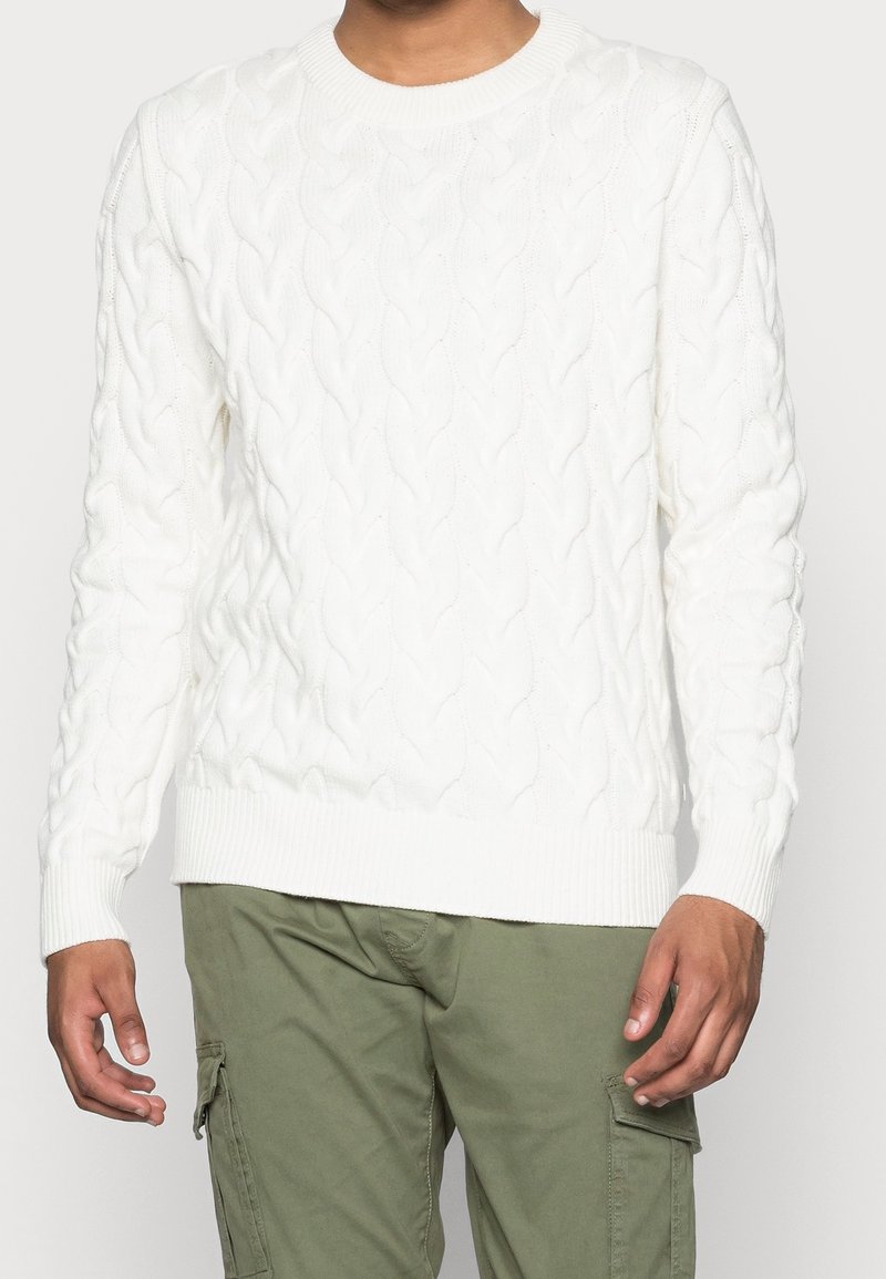 Vit kabelstickad tröja med ribbad rundhals och nederkant. Texturen har sammanflätade mönster. Bärs över olivgröna cargo-byxor.