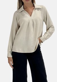Hellbeiges Langarmhemd mit V-Ausschnitt und Kragen, mit einem plissierten Design. Getragen zu dunklen Hosen, die eine lockere Passform zeigen.