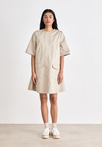 Vestido de algodão bege com mangas curtas, decote redondo, dois bolsos frontais e detalhes em botão nos ombros, apresentando uma silhueta evasê.