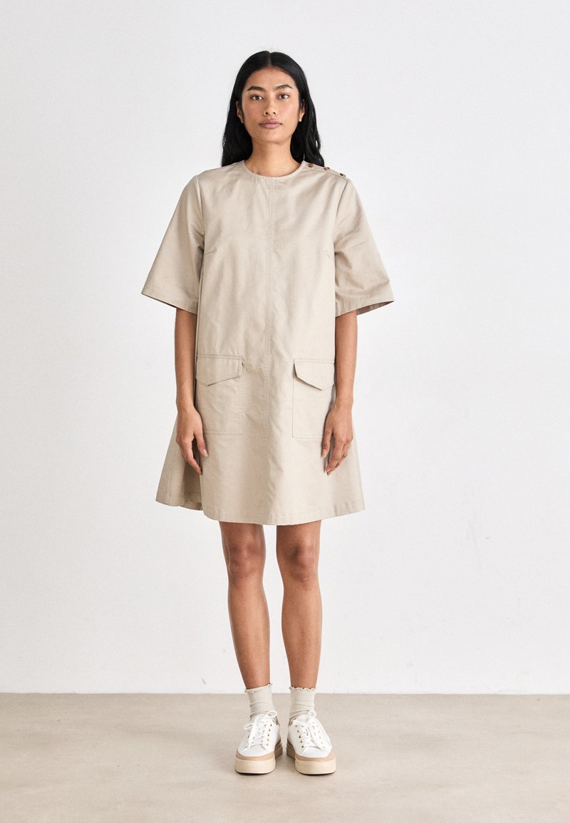 Vestido de algodão bege com mangas curtas, decote redondo, dois bolsos frontais e detalhes em botão nos ombros, apresentando uma silhueta evasê.