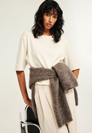 Woman with wavy black hair wears a cream shirt and trousers, with a brown knit sweater tied around her waist, standing by a black chair.