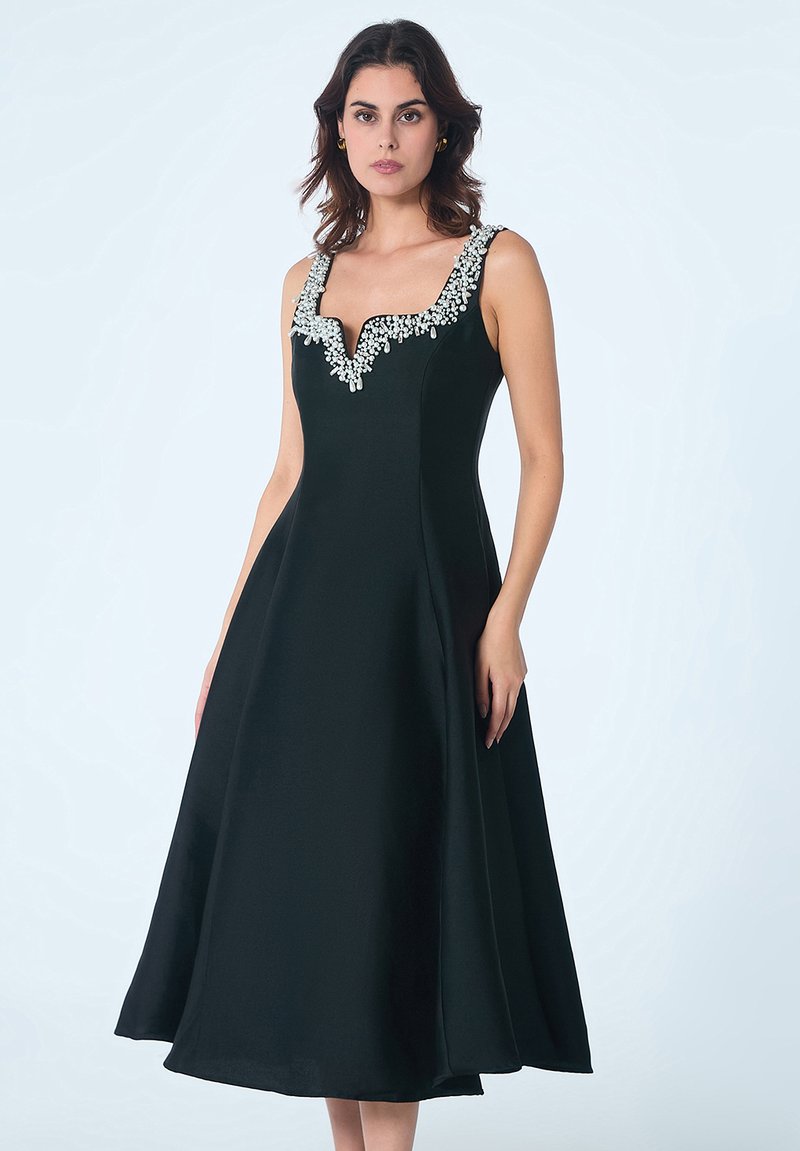 Robe noire en A avec un décolleté en V ornée de détails en perles au niveau du col ; tissu lisse, sans manches et jupe évasée.