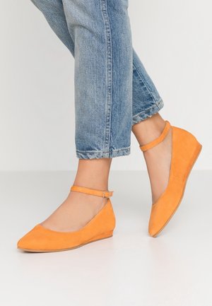 Person, der bærer lyseblå jeans og orange ruskind kilehæle med ankelremme, står på en hvid gulv mod en lys baggrund.