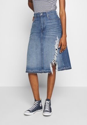 Frau trägt einen knielangen blauen Jeansrock mit ausgefranstem Seitenschlitz, gestreiftes ärmelloses Top und marineblaue High-Top-Sneakers.
