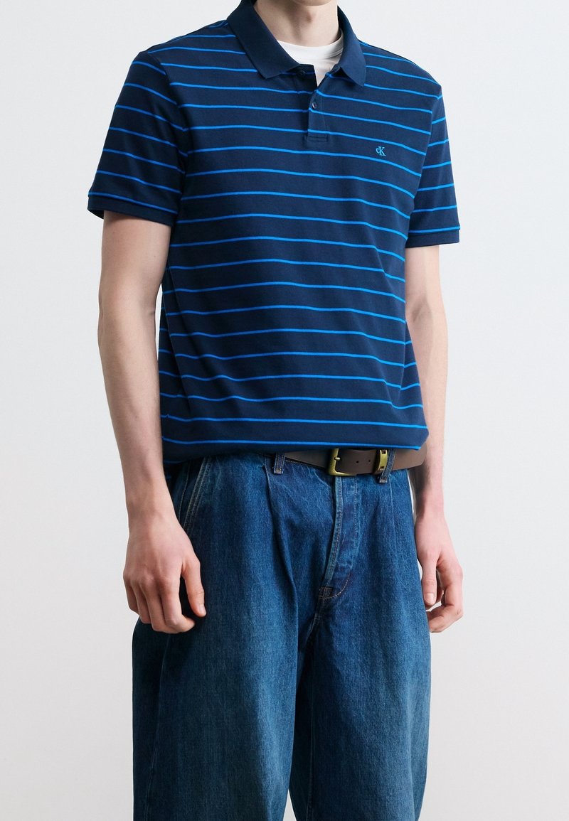 Homme portant un polo bleu foncé avec des rayures horizontales bleu clair et un jean bleu ample avec une ceinture marron sur un fond uni.